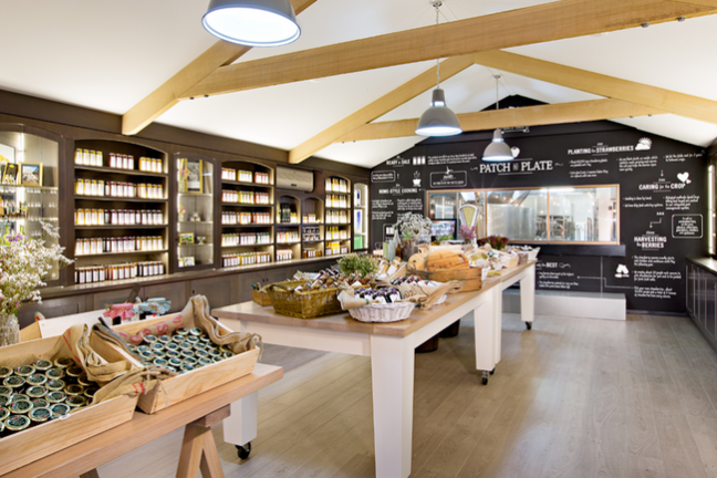 Well-lit farm shop with wooden beams, shelves of jars, and tables displaying various foods.