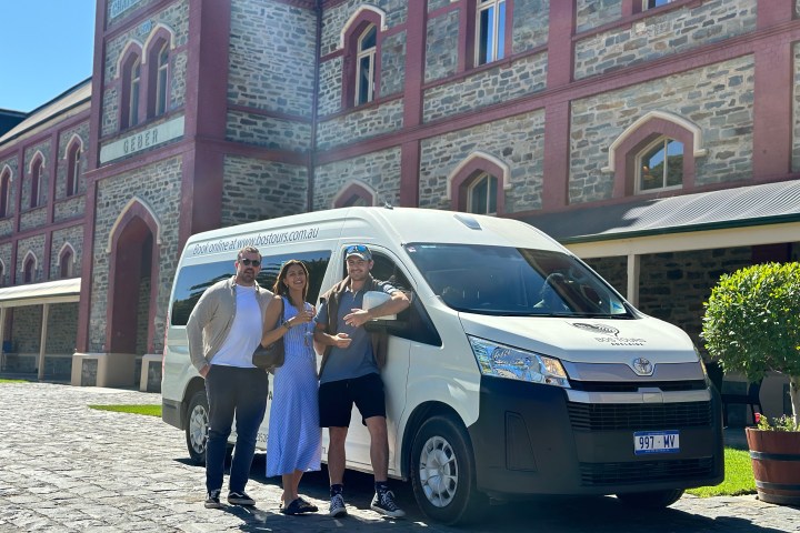 Three people posing in front of a tour van near a brick building.