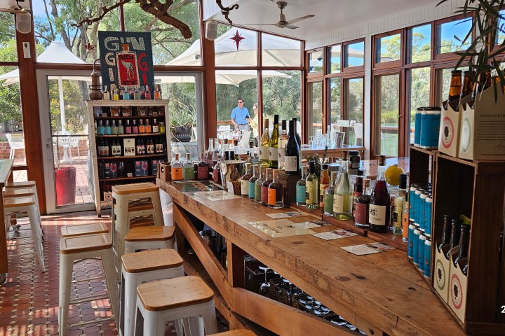 Bar with bottles, stools, and tables beneath a wooden pergola on a sunny day.