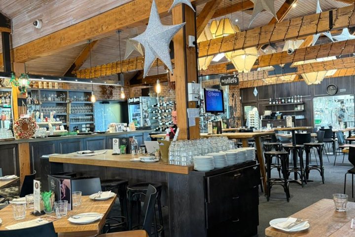 Cozy restaurant interior with wooden beams, star decorations, tables set with plates, and stocked bar.