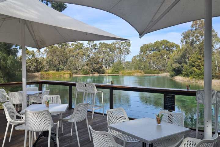 Outdoor cafe with white chairs and tables by a lake under large umbrellas.