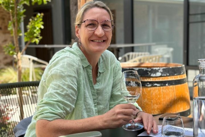 Person in light green shirt smiling and holding a wine glass at an outdoor table.