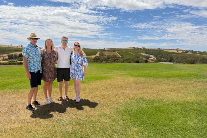 Four people standing on grass with a vineyard and blue sky in the background.