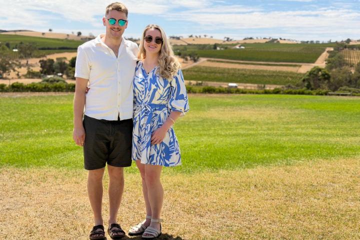 Couple posing in sunglasses on a grassy field with hills and blue sky in the background.
