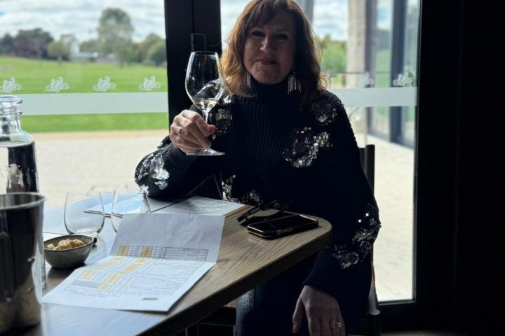 Woman sitting at a table with a wine glass, large windows with a view of grass and sky in the background.