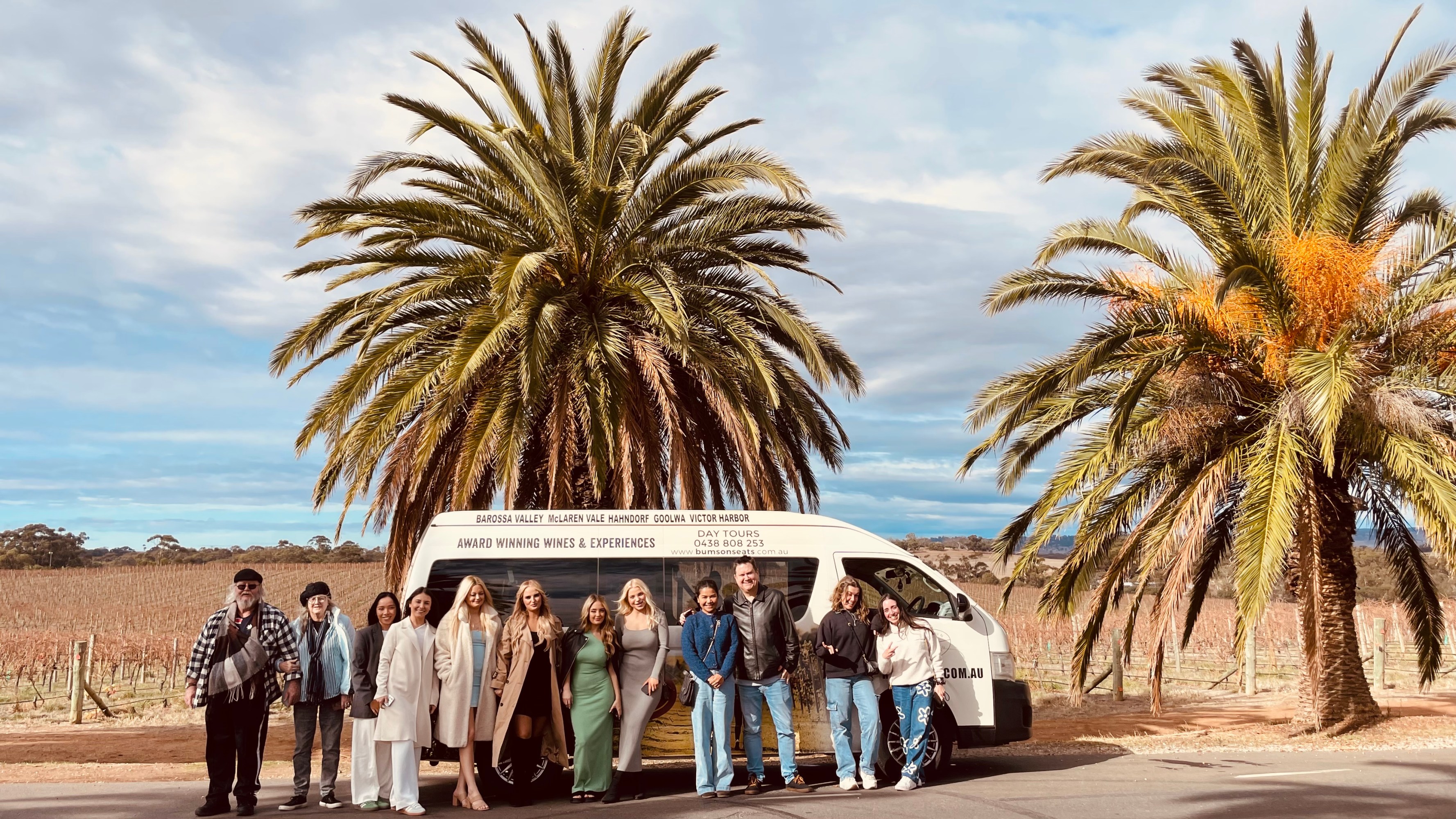 Group of people standing in front of a van and palm trees in a vineyard.