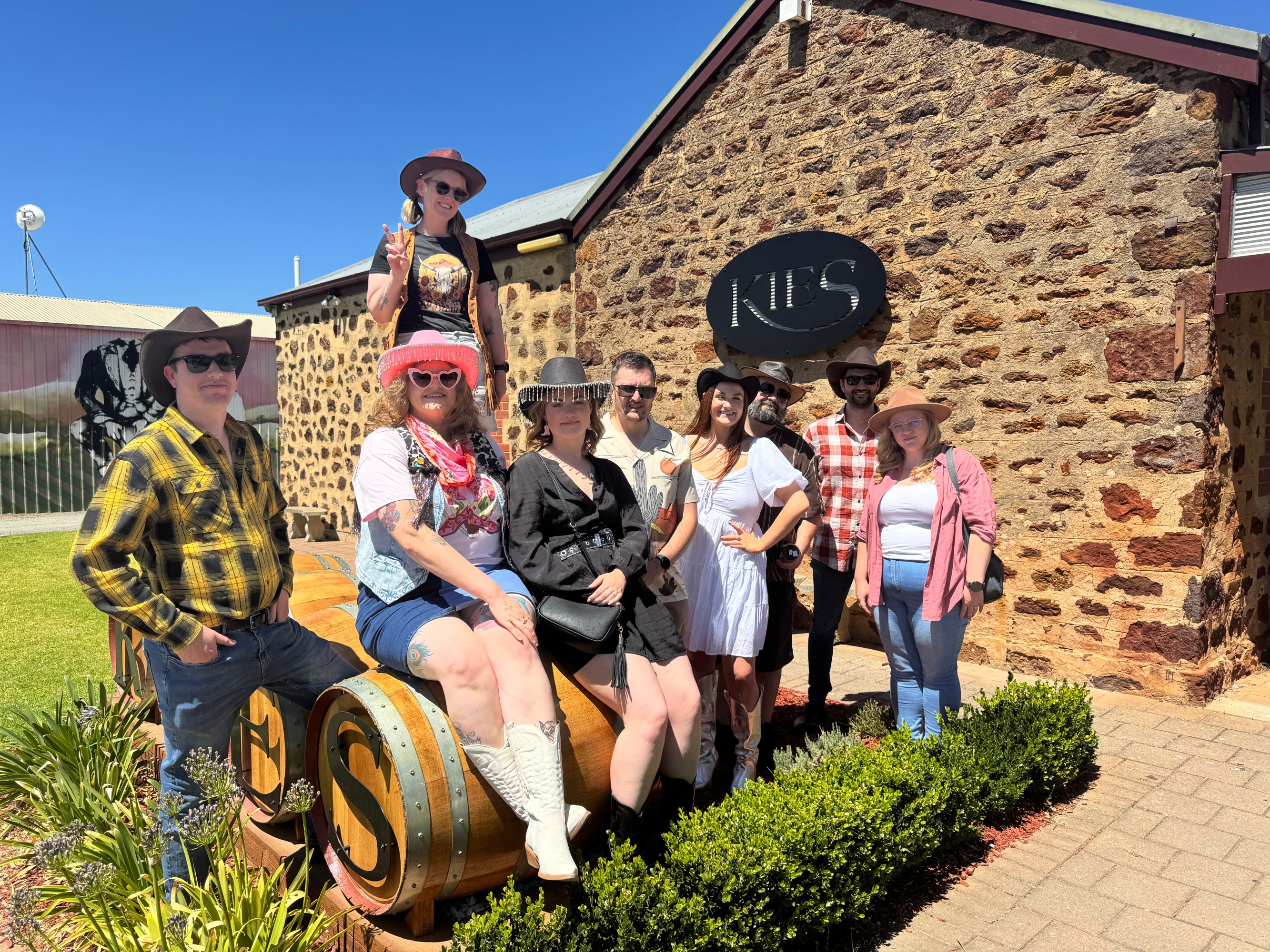 Group of people in casual attire pose in front of a stone building with 'Kies' sign.