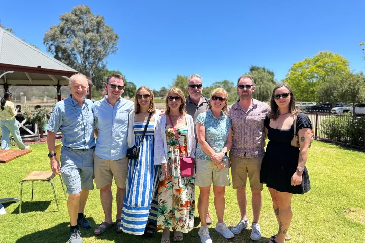 Group of seven people posing outdoors on a sunny day with blue sky and trees in the background.