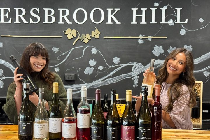 Two women smile behind a bar displaying various wine bottles.