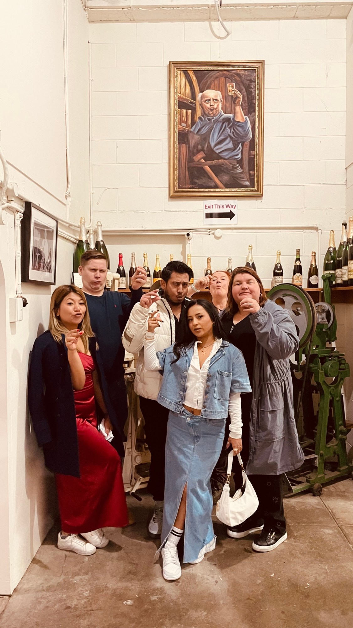 Group of six people posing with drinks in a room, surrounded by champagne bottles and a painting on the wall.