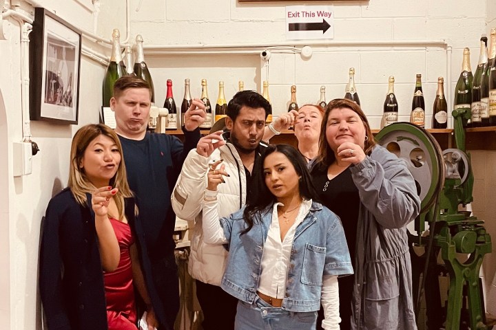 Group of six people posing with drinks in a room, surrounded by champagne bottles and a painting on the wall.
