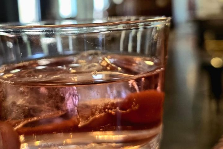 Close-up of a hand holding a glass with a pink drink and ice cubes in a dimly lit room.