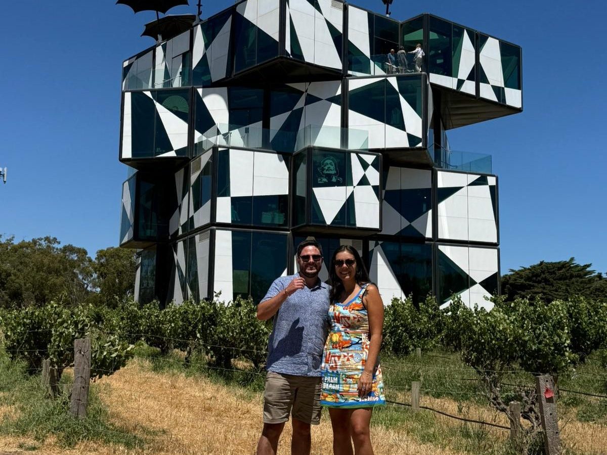 Couple posing in front of a modern, geometric building with umbrellas on top, under a clear blue sky.