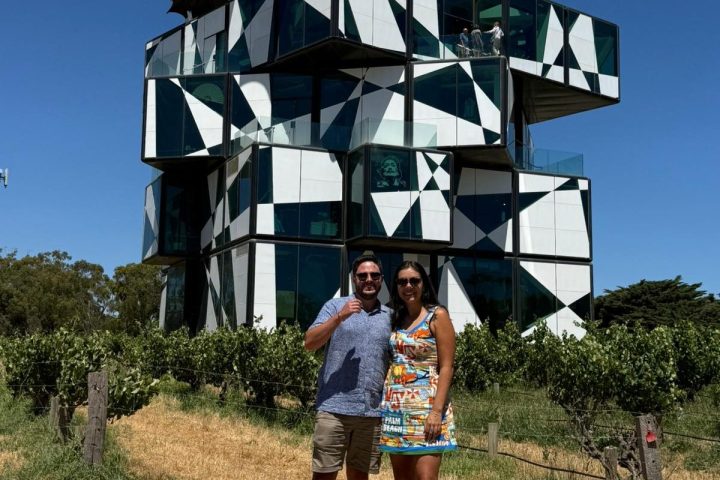 Couple posing in front of a modern, geometric building with umbrellas on top, under a clear blue sky.
