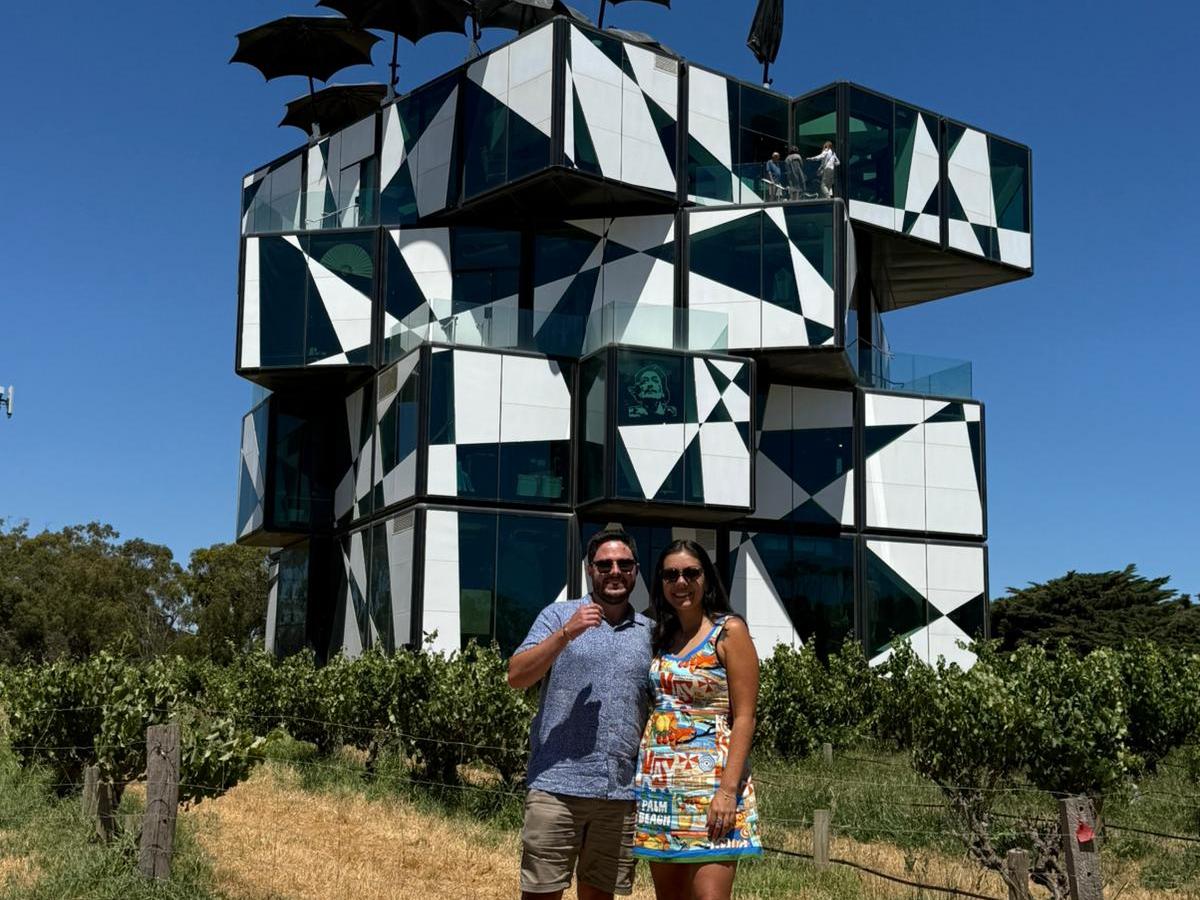 Couple posing in front of a modern, geometric building with umbrellas on top, under a clear blue sky.