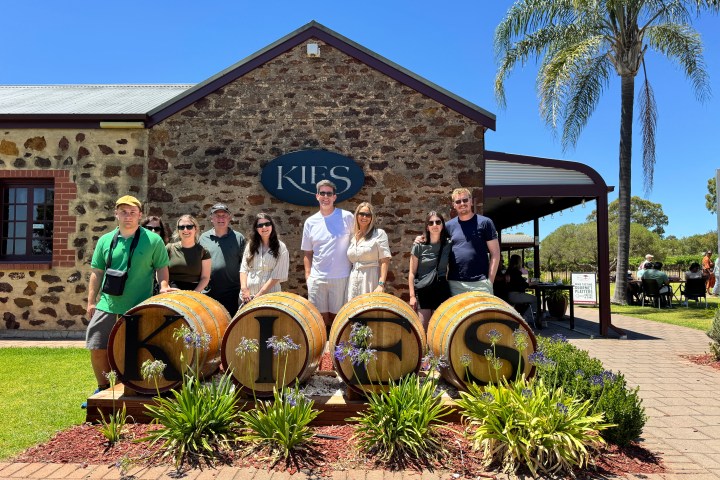 Group of seven people smiling in front of Kies winery barrels and stone building.