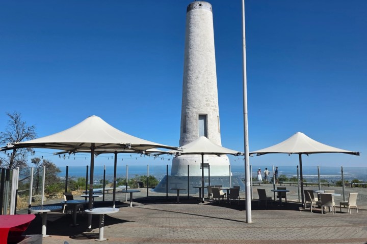 Tall white lighthouse with umbrellas and seating area, clear blue sky.
